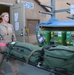 A female military or medical personnel member in tan/khaki flight suit is transporting equipment on a wheeled cart outside a building marked '5318A'. She is pulling a litter/stretcher loaded with olive drab duffel bags, with green medical or oxygen containers visible nearby on a separate cart. A sign reading 'Outbound PT LOX For Mission' is visible in the upper right, indicating an aeromedical evacuation or patient transport staging area.