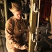 A female military service member wearing a tan/desert flight suit is kneeling inside what appears to be a military cargo aircraft, working on litter stanchion hardware. She is focused on securing or adjusting metal locking mechanisms on a vertical support rail, consistent with aeromedical evacuation equipment setup. The interior environment shows typical military transport aircraft features including cargo floor tracks and safety equipment.