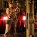 A military aircrew member in tan/khaki flight suit is working inside what appears to be a military cargo aircraft, likely a C-130 or similar transport. The individual is manipulating cargo restraint straps or tie-down equipment, illuminated by red interior lighting. The aircraft interior shows typical military transport features including red troop seats, cargo rails, and safety markings.