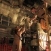 Two military personnel in tan/khaki flight suits are working inside the cargo hold of a large military aircraft, likely a C-130 or similar transport. One person is positioned elevated near the ceiling/upper structure while the other stands below, both appearing to pass or handle equipment between them. The interior shows characteristic military aircraft features including insulated padding, red cargo netting, and exposed mechanical systems.