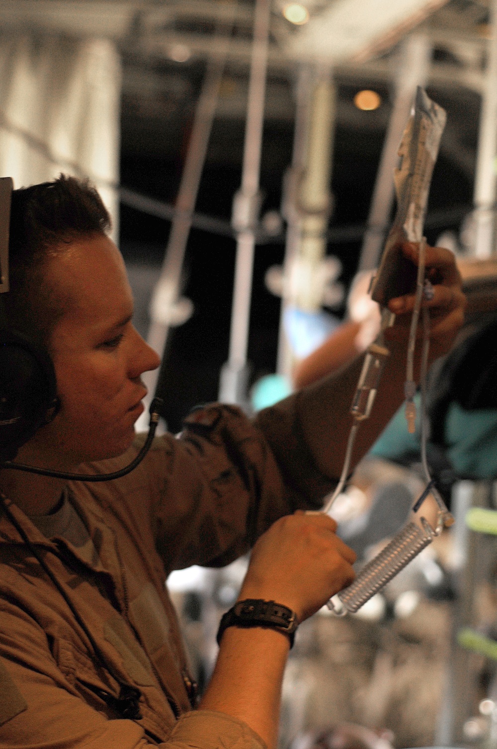 A military medical personnel, wearing a tan/olive flight suit and communications headset, is handling an IV bag and tubing inside what appears to be a military aircraft configured for aeromedical evacuation. The individual is focused on preparing or adjusting intravenous medical equipment, with IV lines and hanging hardware visible overhead. The setting suggests an in-flight medical care environment typical of military casualty transport operations.