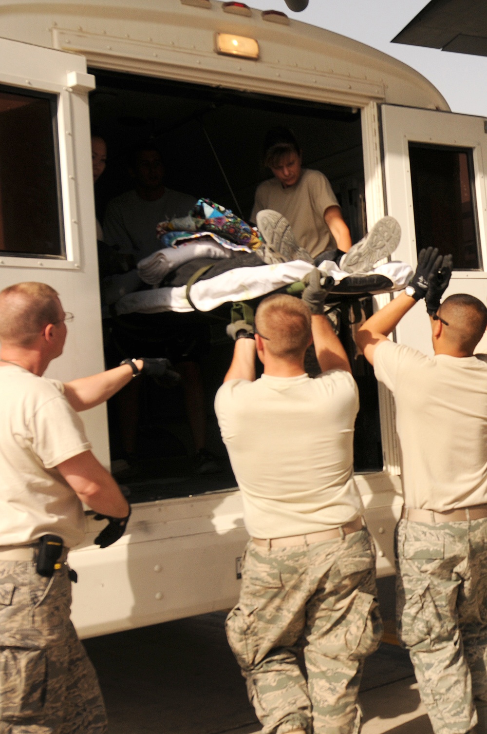Military personnel in US Air Force uniforms (ABU/ACU pattern) are loading or unloading a patient on a stretcher/litter through the open side door of a white bus or transport vehicle. At least three uniformed service members outside are lifting the stretcher upward while individuals inside the vehicle assist in receiving it. The scene appears to be a medical evacuation or patient transfer operation.