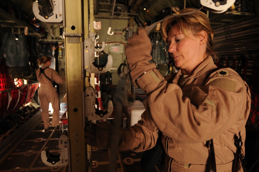 Two female military personnel in tan/desert flight suits are working inside what appears to be a military cargo aircraft, likely a C-130 or similar transport. The foreground subject is inspecting or adjusting mechanical hardware on a vertical track or rail system, while a second person works in the background near the aircraft's rear. The interior shows typical military cargo aircraft features including ribbed walls, tie-down tracks, and equipment.