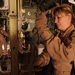 Two female military personnel in tan/desert flight suits are working inside what appears to be a military cargo aircraft, likely a C-130 or similar transport. The foreground subject is inspecting or adjusting mechanical hardware on a vertical track or rail system, while a second person works in the background near the aircraft's rear. The interior shows typical military cargo aircraft features including ribbed walls, tie-down tracks, and equipment.