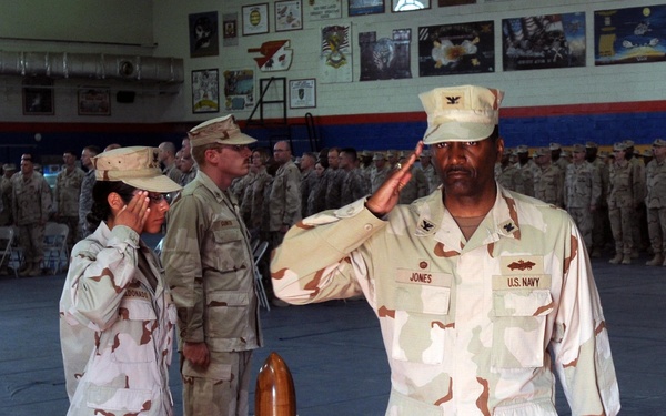 A formal U.S. military ceremony taking place inside what appears to be a gymnasium or drill hall. A U.S. Navy officer named 'Jones,' wearing the rank of Captain (O-6, indicated by the eagle insignia on his cover), is rendering a salute while walking on a red carpet. Multiple service members in desert camouflage uniforms (DCUs) are standing at attention in formation in the background.