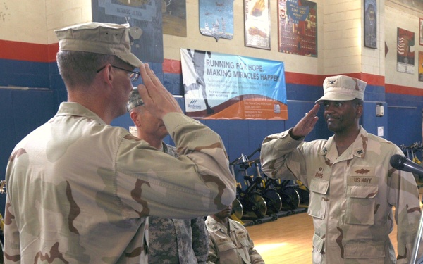 A military ceremony is taking place inside what appears to be a gymnasium or fitness facility. Two service members in desert camouflage uniforms are exchanging salutes, with one identified as U.S. Navy personnel (name tag reads 'JONES') standing near a podium with a microphone. Several other military personnel in various camouflage uniforms are seated in the background.