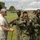 The image shows a military training exercise taking place on an outdoor field. A trainer in US Air Force digital camouflage (ABU) pattern trousers and a tan t-shirt is observing several soldiers in woodland camouflage uniforms who are practicing weapons handling with AK-pattern rifles. A second trainer in military uniform is visible in the background.