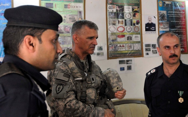 The image shows a U.S. Army soldier in ACU (Army Combat Uniform) with Airborne and 25th Infantry Division patches, standing alongside two individuals in dark police/security uniforms. The setting appears to be an indoor briefing or meeting room with Arabic-language informational posters on the wall. This appears to be a military-police coordination or advisory meeting.