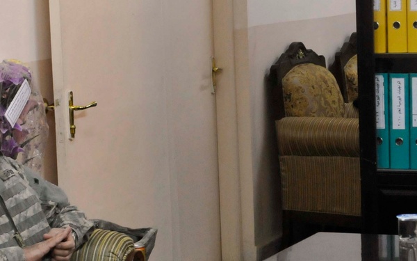 The image shows a meeting between two U.S. Army soldiers in combat uniforms and a local official seated behind a desk with an Arabic nameplate. The setting appears to be an official office, likely in Iraq or Afghanistan, with yellow binders on shelves in the background. This appears to be a liaison or coordination meeting between U.S. military personnel and a local security or police official.