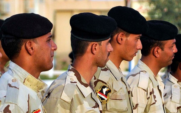 A formation of military personnel standing in a line, wearing desert camouflage uniforms and black berets. Several soldiers display shoulder patches including what appears to be the Iraqi flag patch and unit insignia. The setting appears to be an outdoor military compound or base.