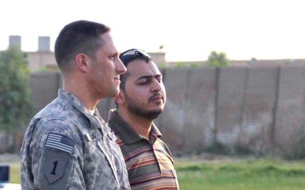 A U.S. Army soldier in ACU (Army Combat Uniform) stands alongside a civilian man at what appears to be an outdoor podium or lectern. The soldier wears a patch indicating the 1st Infantry Division ('Big Red One') on his left shoulder, along with a U.S. flag patch. The setting appears to be a forward operating base or similar military compound, based on the concrete blast walls visible in the background.