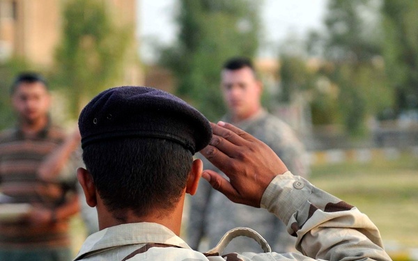 A soldier wearing a desert three-color camouflage uniform and dark beret is photographed from behind while rendering a salute. In the background, at least two other individuals are visible, one appearing to wear a different military uniform. The setting appears to be an outdoor area with grass and trees.