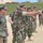 A group of military personnel in woodland camouflage (BDU) uniforms are lined up on a sandy outdoor range area. One individual in the foreground is extending his arm in a pointing or directing gesture, while others stand at attention or in relaxed stances. The setting appears to be a training exercise or range qualification event near a coastal or sandy terrain.