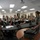 A military briefing or training session is taking place in a classroom setting, with approximately 25 uniformed personnel seated around tables and standing along the back wall. A female soldier in camouflage uniform is standing at the front, apparently leading or instructing the group. The room contains military equipment including a field radio/communications device on a nearby table.
