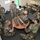 A group of military personnel in woodland camouflage uniforms are gathered in a classroom setting for what appears to be a training session. Several soldiers are clustered around a round table examining military communications equipment, while others are seated at desks in the background. The scene depicts hands-on technical instruction with a piece of field radio or communications gear as the focal point.
