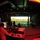 A darkened production or simulation control room captured with a fisheye lens, showing a large projection screen displaying a desert/arid landscape scene. A technician or operator sits at a workstation with a computer monitor in the foreground, while several silhouetted individuals are positioned near the projection screen in the background. The space appears to be a professional theater, simulation facility, or film production stage with red ambient lighting and exposed ceiling infrastructure.