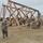 Military personnel in camouflage uniforms and hard hats are constructing a large wooden truss structure on an open dirt field. The structure appears to be a bridge or roof truss framework assembled from heavy timber lumber. A military truck is visible in the background, suggesting this is likely a combat engineering or construction training exercise.