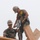 Two workers in military-style camouflage uniforms and tan hard hats are working at elevation on a wooden roof frame structure. The standing worker is operating a large power drill or auger into a wooden beam while the other worker assists and stabilizes. Both are equipped with safety harnesses and fall protection equipment.