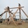 Military personnel in camouflage uniforms are constructing a large wooden timber-frame structure on a dirt field. A military-green telescopic forklift/telehandler is being used to assist in lifting and positioning heavy wooden beams. The construction appears to be a training exercise at a military installation.
