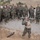 A military training exercise is taking place outdoors on a gravel range area. One soldier in the center is demonstrating the proper shouldering technique of a rocket launcher (appears to be an AT4 or similar anti-tank weapon) to a large group of approximately 25-30 soldiers standing in a semicircle. The soldiers are wearing woodland camouflage BDUs, suggesting this is a pre-2005 era U.S. military training event.