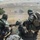 Three soldiers in woodland camouflage uniforms are positioned on elevated ground overlooking an open, arid landscape. They appear to be engaged in a live-fire exercise or combat operation, with spent brass casings visible on the ground. Smoke or dust is visible in the distance, suggesting active firing downrange.