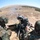 Two soldiers in woodland camouflage uniforms are operating what appears to be an automatic grenade launcher from an elevated firing position. The weapon has been fired, as evidenced by visible smoke near the muzzle and spent casings/rounds visible in an ammunition box. The image is taken with a wide-angle fisheye lens, showing an arid, open landscape in the background.