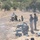 A military firing range exercise showing multiple personnel in camouflage uniforms operating automatic grenade launchers or heavy machine guns in a line formation. Several soldiers are in firing positions while two individuals in mixed civilian/military attire and helmets appear to be observing or instructing. Spent casings and ammunition boxes are scattered across the dry, arid terrain.