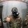 Three U.S. Navy personnel wearing woodland camouflage BDUs and M40-series gas masks are photographed inside what appears to be a gas chamber training facility. The central figure, identified by name tape as 'THURSBY,' is giving a thumbs-up gesture toward the camera. The image is captured with a fisheye lens, creating a wide-angle distortion effect.