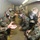A group of military personnel wearing gas masks and woodland camouflage uniforms are inside a narrow enclosed corridor, likely a gas chamber training facility. The individuals are arranged in two lines facing each other, with some removing or holding their gas masks, consistent with standard CBRN (Chemical, Biological, Radiological, Nuclear) training exercises. A hazy atmosphere is visible in the corridor, suggesting the presence of CS gas or a similar training agent.