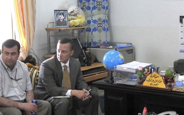 A meeting is taking place in what appears to be an official office, likely in Iraq or another Middle Eastern country. Four men are present: one in US military uniform, two in civilian attire seated on a sofa, and one man seated behind a large desk who appears to be a local official. Arabic text is visible on a nameplate on the desk, and an Iraqi flag is displayed.