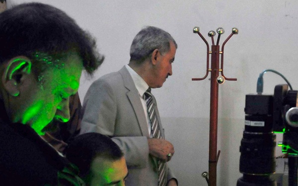 Three men are gathered around a workstation illuminated by intense green laser light, suggesting a laser optics or holography laboratory setting. One man is seated and operating equipment while two others observe. The green light dominates the scene, casting strong colored shadows across all subjects and surfaces.