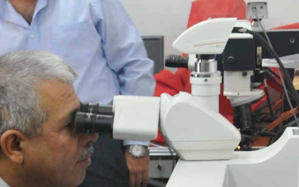 A middle-aged man wearing a dark suit and tie is looking through the eyepiece of a large, modern research microscope in what appears to be a laboratory setting. A second person in a light blue shirt is standing in the background. The lab contains multiple microscopes, computers, and associated equipment.