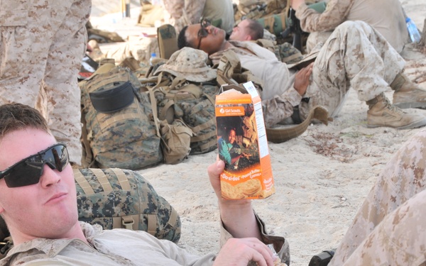 A U.S. Marine in desert MARPAT camouflage is reclining on the ground during a rest period, holding up a box of Girl Scout cookies (appears to be Do-si-dos/Peanut Butter Sandwich cookies). Several other Marines are visible in the background, also resting with tactical gear and backpacks scattered around them. The setting appears to be an arid, sandy outdoor environment.