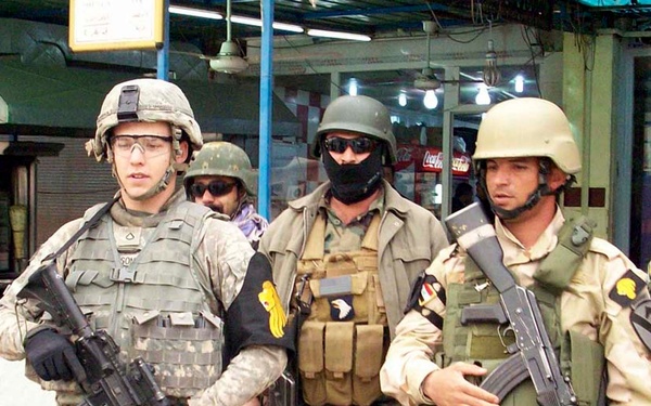 The image shows a group of armed soldiers conducting what appears to be a patrol or security operation in an urban environment. At least three soldiers are visible in the foreground, wearing a mix of U.S. military and other military uniforms with body armor and helmets. The setting appears to be a commercial street or marketplace.