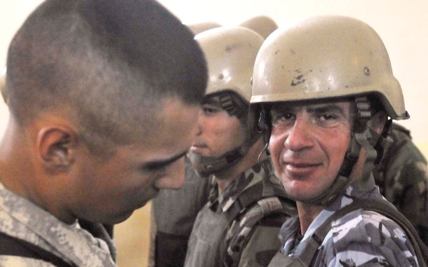 A U.S. Army soldier wearing 1st Cavalry Division insignia and an American flag patch is affixing or examining a yellow lion patch on the sleeve of a local security force member wearing blue-gray camouflage uniform and a tan helmet. Additional personnel in similar local force uniforms and helmets are visible in the background. The scene appears to depict a military advisory or training interaction.