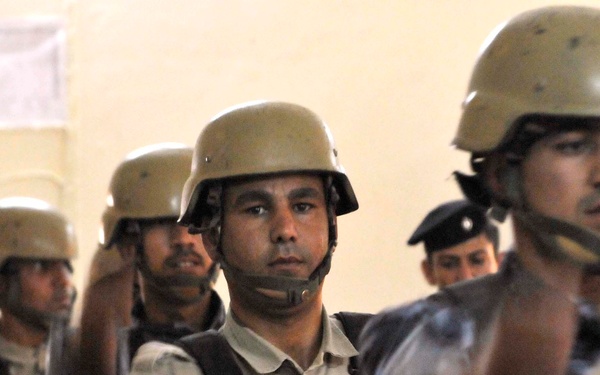 The image shows a group of military or security personnel wearing tan/olive combat helmets and body armor vests. They appear to be gathered indoors, possibly during a briefing or assembly. The personnel are wearing a mix of tactical gear including plate carriers and appear to be in a formal formation or group setting.