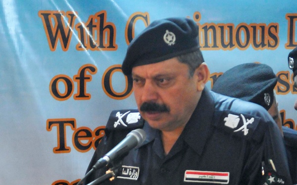 A senior Iraqi police or interior ministry official in black uniform is speaking at a podium with a microphone. The podium displays an official seal of what appears to be the Iraqi Ministry of Interior. A banner in English and Arabic is visible in the background.