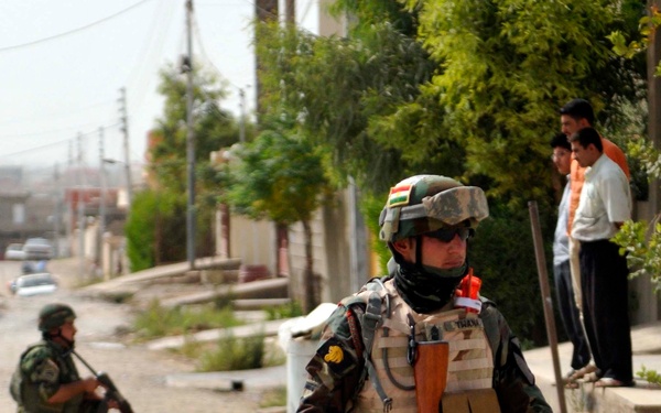 Two armed soldiers in camouflage uniforms conduct a patrol or security operation on a residential street. The foreground soldier wears a helmet with a small flag patch, tactical vest, sunglasses, and carries an AK-pattern rifle. Civilians are visible observing from the sidewalk in the background.