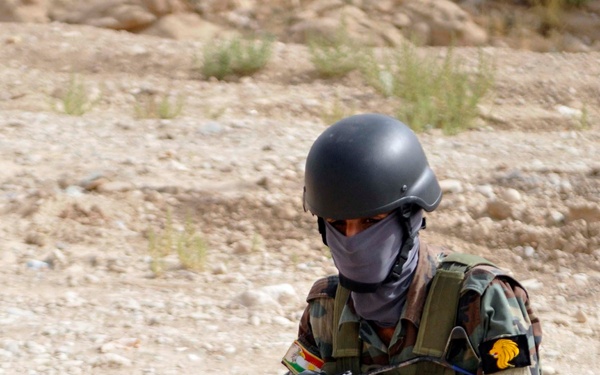 A masked and helmeted soldier in woodland camouflage uniform is kneeling in a low-ready position on rocky, arid terrain. The individual is equipped with an AK-pattern rifle, a tactical vest with pouches, and a face covering. Visible patches on the uniform suggest affiliation with a Kurdish military force.