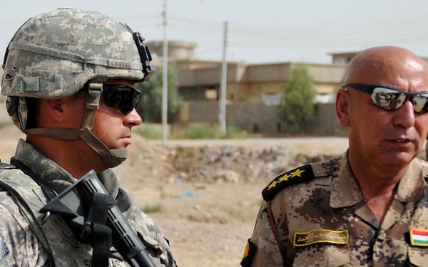 Two military personnel are standing together in what appears to be an arid, urban environment. The soldier on the left wears U.S. Army UCP camouflage with a 10th Mountain Division patch and carries an M4-style rifle, while the officer on the right wears Kurdish Peshmerga uniform with three-star rank insignia and a Kurdistan flag patch. A third soldier is visible in the background near a building.