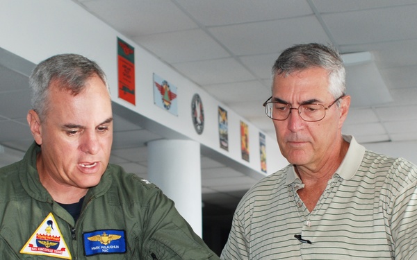 A military officer in an olive green flight suit is reviewing what appears to be a map or chart with a civilian man wearing a striped polo shirt. They are leaning over a glass-topped table in what appears to be a military facility or briefing room. Aviation-related posters and insignia are visible on the walls in the background.