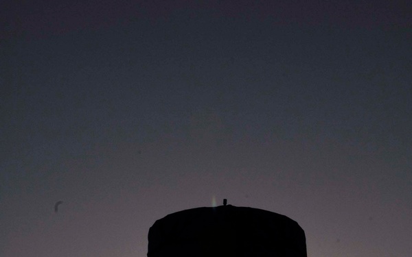A dramatic silhouette photograph taken at sunset showing two individuals working with a large cylindrical piece of equipment mounted on a tripod. The backlit composition places the sun directly behind the tripod structure, creating a strong contrast between the dark figures and the warm gradient sky. The subjects appear to be in military or technical field gear based on their clothing silhouettes.