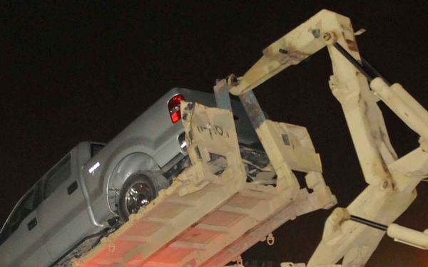 A nighttime scene showing a gray civilian pickup truck being loaded onto a military flatbed recovery trailer using a hydraulic crane/loader arm. The recovery vehicle appears to be a military-grade heavy equipment transporter painted in desert tan/sand color. The operation is taking place on what appears to be a military installation or forward operating base.