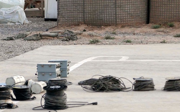 The image shows a collection of surveillance and security equipment laid out on a concrete surface, likely for inventory, inspection, or deployment purposes. Multiple security cameras, coiled cables, metal enclosures, and electronic equipment are organized in rows. The setting appears to be an outdoor compound or base facility.