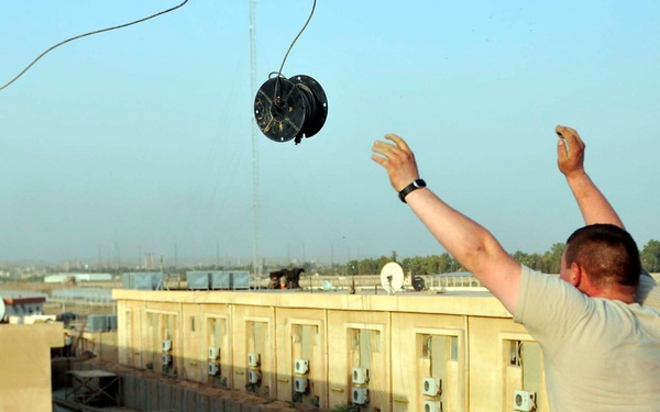 A soldier in US Army ACU (Army Combat Uniform) is standing on a rooftop and throwing a cable spool or wire reel into the air. The setting appears to be a forward operating base (FOB) or military installation in a Middle Eastern environment, characterized by tan/beige modular buildings with multiple air conditioning units. The action suggests the soldier is deploying or running communications cable between buildings.