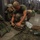 A male service member wearing a green t-shirt and camouflage trousers is crouched on a concrete floor organizing or packing military gear and equipment. The setting appears to be an indoor military storage or staging area with metal cage lockers lining the walls. Multiple pieces of tactical equipment, packs, and gear are spread across the floor around him.