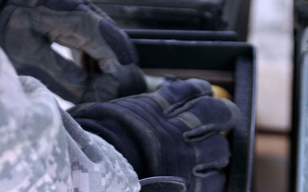 A gloved hand in tactical/military gloves is handling a row of 40mm grenade rounds stored in a foam-lined ammunition case. The rounds display characteristic dual-section construction with brass-colored ogive tips and dark steel bodies. The person is wearing US Army ACU (Army Combat Uniform) digital camouflage pattern.