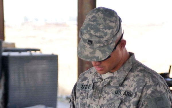 A U.S. Army soldier identified by name tape as 'Smith' is standing at a work surface, examining or handling an item while wearing ACU (Army Combat Uniform) and tactical gloves. The soldier displays a Staff Sergeant rank insignia on the chest and a '1' patch on the left sleeve, consistent with the 1st Infantry Division. Various military equipment and supplies are arranged on the surface in front of him.