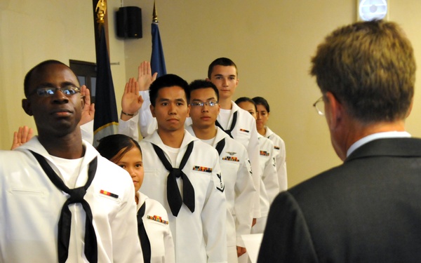 A group of U.S. Navy personnel in white dress uniforms are taking an oath, with their right hands raised, before a civilian official in a dark suit who faces them with his hand raised. The ceremony appears to be a formal military re-enlistment or swearing-in ceremony conducted indoors. Multiple sailors of diverse backgrounds are visible standing in formation.