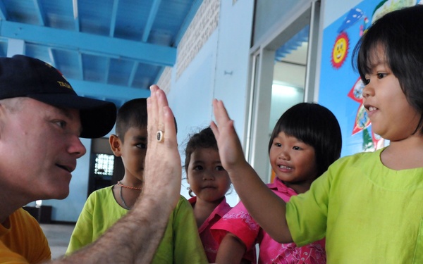 An adult male wearing a yellow t-shirt and dark baseball cap is giving high-fives to a group of young children at what appears to be a school or community center. The children are wearing bright green and pink uniforms or smocks, suggesting an educational or institutional setting. The interaction appears cheerful and friendly.